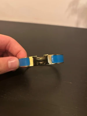 Blått armband med gulddetaljer - Snyggt armband i metall med guldfärgad finish och blå emalj. Armbandet har en tydlig H-detalj framtill som ger en exklusiv känsla. Perfekt accessoar för att lyfta din outfit med en färgklick och stilren design.