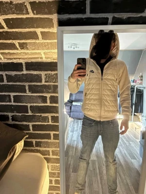 Vit quiltad hoodiejacka från Nike - Snygg vit hoodiejacka från Nike med quiltad front och mjuka ärmar. Jackan har dragkedja, huva med dragsko och Nike-logga på bröstet. Perfekt för dig som vill ha en sportig och stilren look. Passar till jeans eller träningskläder.