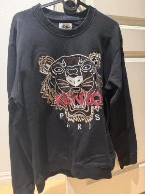 Svart Kenzo tröja med tigertryck - Original pris 2900! Svart tröja från Kenzo med ikoniskt broderat tigerhuvud och texten 'KENZO PARIS' framtill. Tröjan har rund halsringning och ribbade muddar. Perfekt statement-plagg för dig som vill sticka ut.