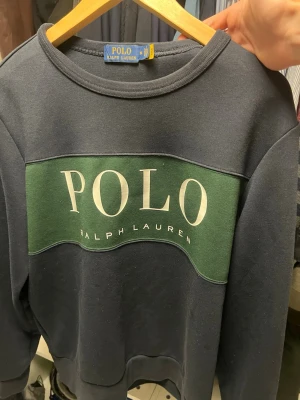 Blå sweatshirt från Polo Ralph Lauren - Mörkblå sweatshirt från Polo Ralph Lauren med grönt parti över bröstet och stor vit logga. Klassisk rund halsringning och ribbade muddar vid ärmslut och nederkant. Perfekt för en avslappnad och stilren look. Kom med prisförslag