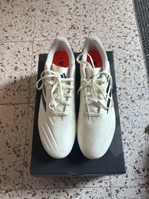 Vita Adidas Copa fotbollsskor - Säljer ett par vita Adidas Copa fotbollsskor med svarta detaljer och röda innersulor. Skorna har klassisk snörning och är designade för bra grepp och kontroll på planen. Perfekta för dig som vill ha stilrena och funktionella skor till match eller träning.