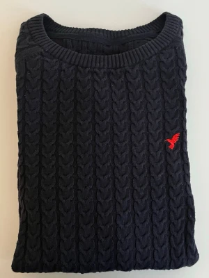 Mörkblå Pier one tröja i storlek XS - Mörkblå kabelstickad tröja med rund halsringning och ribbade muddar. Tröjan har en liten röd broderad logga på bröstet och är tillverkad i ett mjukt stickat material. Perfekt för dig som gillar klassisk och stilren look.