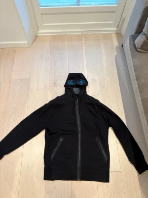 C.P Company Hoodie - Säljer en svart hoodie med dragkedja och två fickor med dragkedja framtill. Huvan har inbyggda glasögon vilket ger en unik och cool look. Perfekt för dig som vill sticka ut. Tillverkad i mjukt sweatshirtmaterial.