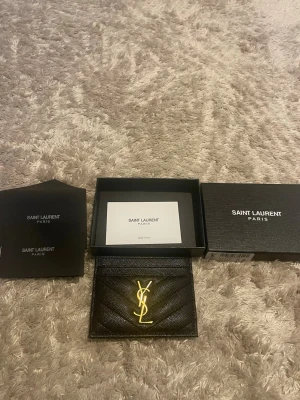 Svart korthållare från Saint Laurent - Snygg svart korthållare i läder från Saint Laurent med guldfärgad YSL-logga framtill. Korthållaren har quiltad struktur och flera fack för kort. Perfekt accessoar för dig som gillar stilrena detaljer och lyxig känsla.