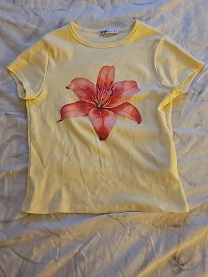 Gul t-shirt med stor blomma - Snygg gul t-shirt med en stor rosa blomma tryckt på framsidan. T-shirten har rund hals och korta ärmar. Perfekt för dig som vill ha en färgklick i garderoben. Tillverkad i mjuk bomull som känns skön mot huden.