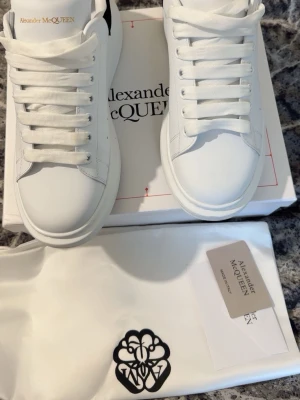 Vita Alexander McQueen sneakers - Stilrena vita sneakers från Alexander McQueen med chunky sula och svart detalj på hälen. Skorna har breda vita skosnören och diskret logga i guld på plösen. Tillverkade i slätt läder för en clean look. Perfekta för dig som gillar minimalistisk och trendig stil. Har använt den två gånger när men den har ingen lukt, skada Elr något liknande. Dem är för små för mig därför tänker sälja dem. 