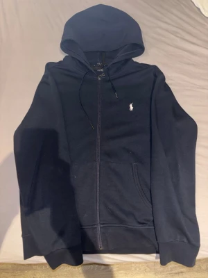 Polo Ralph tröja - Snygg marinblå hoodie från Polo Ralph Lauren med dragkedja framtill och vit broderad logga på bröstet. Tröjan har huva med dragsko, två fickor och ribbade muddar. Perfekt för en avslappnad och stilren look.