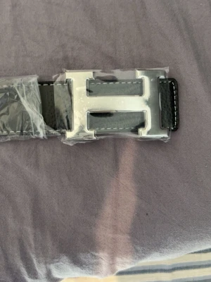 Svart bälte  - Snyggt svart bälte med ett stort, silverfärgat H-format spänne. Bältet har vita sömmar längs kanterna och är gjort i skinn. Perfekt accessoar för att lyfta din outfit med en stilren detalj.
