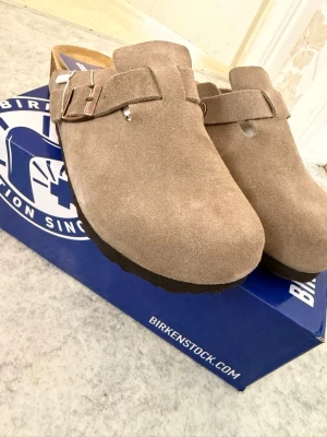 Beige mocka tofflor från Birkenstock - Säljer ett par klassiska beige tofflor från Birkenstock i mjuk mocka. Modellen har en justerbar rem med metallspänne och en bekväm korksula. Perfekta för dig som vill ha både stil och komfort i vardagen. !!SKICKAR ALLTID INOM 24H** **PRIS KAN DISKUTERAS** 
