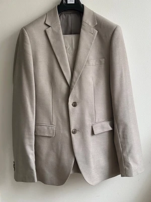 Beige kostym av märket Riley från Brothers - Klassisk beige kostym av märket Riley med två knappar framtill, smala slag och tre fickor. Kavajen har en slät och stilren design med helfodrad insida. Har endast använt den en gång och är numera för liten för mig, kvitto finns