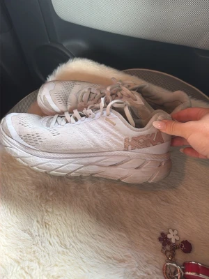 Vita Hoka sneakers med tjock sula - Säljer ett par vita Hoka sneakers med tjock, dämpad sula och ventilerande mesh-ovandel. Skorna har klassisk snörning och diskret Hoka-logga på sidan. Perfekta för dig som gillar sportig och clean stil.
