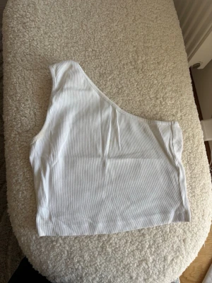 Vit ribbad one shoulder topp - Säljer en vit ribbad crop top med one shoulder-design. Toppen har smala ribbor och är croppad vilket ger en trendig look. Perfekt för dig som vill ha något stilrent men ändå unikt.
