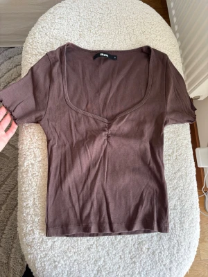 Brun ribbad topp från Bik Bok - Snygg brun ribbad topp från Bik Bok med korta ärmar och djup v-ringning. Toppen har en liten rynkdetalj framtill och är tillverkad i ett mjukt, stretchigt material som sitter bekvämt på kroppen. Perfekt att styla med jeans eller kjol.