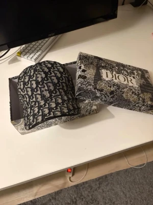 Monogram keps från Dior - Säljer en snygg keps från Dior med klassiskt monogrammönster i svart och grått. Kepsen är tillverkad i 100% bomull och har justerbar rem bak. Kommer i originalbox med exklusivt mönster. Perfekt för dig som gillar lyxiga accessoarer.