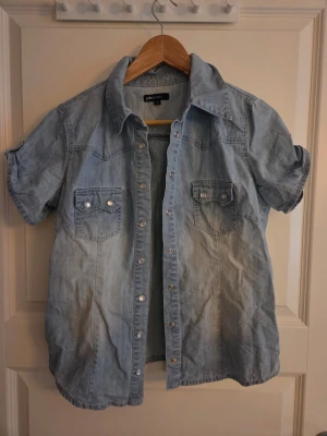 2000s Ljusblå jeansskjorta  - Säljer en ljusblå jeansskjorta från jsfn denim i storlek 40 men passar mindre.. Skjortan har korta ärmar, tryckknappar framtill och två bröstfickor med lock. Perfekt för dig som gillar en avslappnad och cool stil.