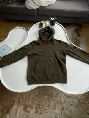 Olivgrön CP company hoodie med huva - Säljer en olivgrön CP company hoodie med huva i mjukt och skönt material. Hoodien har ribbade muddar och den ikoniska patchen på vänster ärm och en klassisk passform som passar perfekt till en avslappnad stil. Materialet känns fleece-liknande och är riktigt bekvämt. Storlek 12 år