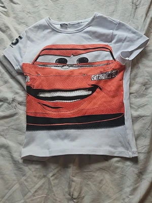 Vit t-shirt med Cars motiv - Säljer en vit t-shirt med stort tryck av Blixten McQueen från Disney Cars på framsidan. T-shirten har korta ärmar och rund hals. Materialet ser ut att vara bomull och trycket är i rött, svart och vitt. Perfekt för dig som gillar animerade filmer och coola motiv.
