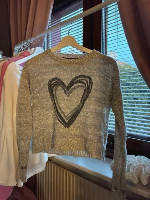 Grå stickad tröja med hjärta KappAhl - Grå stickad tröja från KappAhl med ett stort grått/svart hjärta på bröstet. Tröjan har lång ärm. Perfekt för dig som gillar en avslappnad och trendig stil.🌸