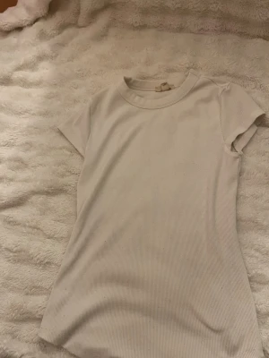Vit ribbad t-shirt från H&M - Säljer en enkel och stilren vit ribbad t-shirt från H&M. T-shirten har korta ärmar, rund halsringning och är gjord i mjuk bomull. Perfekt basplagg till garderoben och lätt att matcha med allt.