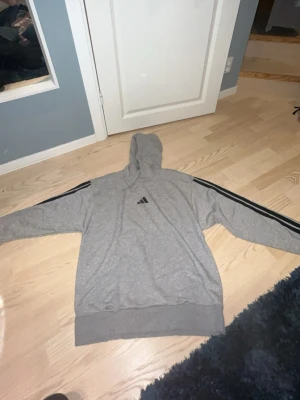 Grå Adidas hoodie med svarta ränder - Snygg grå hoodie från Adidas med klassiska svarta ränder längs ärmarna och Adidas-logga på bröstet. Hoodien har en luva och breda muddar vid ärmslut och nederkant. Perfekt för en avslappnad och sportig stil.