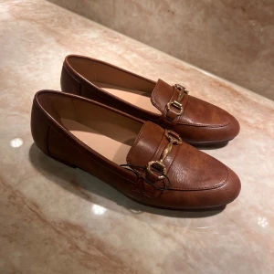 Bruna loafers med gulddetaljer - Snygga bruna loafers i läderimitation med guldfärgade metallspännen över vristen. Skorna har en klassisk, rundad tå och låg klack. Perfekta för dig som gillar stilrena och tidlösa skor med en touch av elegans.