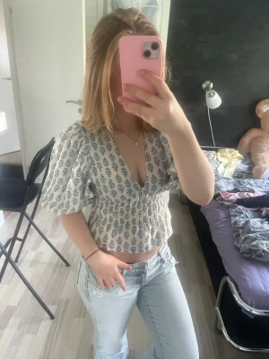 Hm topp - Supersöt vit blus med blått blommigt mönster och puffiga korta ärmar. Djup v-ringning och smockad midja som ger en feminin siluett. Perfekt till jeans eller kjol för en trendig look.