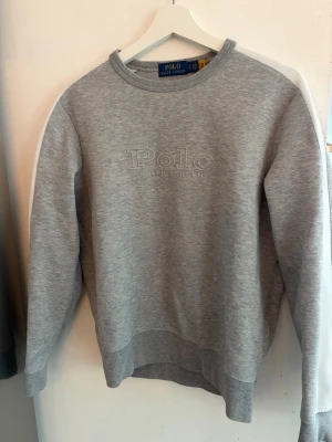 Grå sweatshirt från Polo Ralph Lauren - Snygg grå sweatshirt från Polo Ralph Lauren med broderad logga framtill. Klassisk rund halsringning och ribbade muddar vid ärmslut och nederkant. Perfekt för en avslappnad och stilren look.