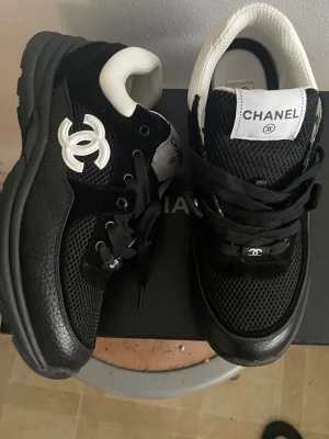Svarta Chanel sneakers med vit logga - Snygga svarta sneakers från Chanel med vit logga på sidan och vit detalj vid hälen. Ovandelen är i mesh och läder, med svarta skosnören och tydlig Chanel-märkning på plösen. Perfekta för en trendig streetstyle-look.