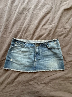 Blå jeanskjol med råa kanter - Trendig blå jeanskjol i kort modell med slitna detaljer och råa kanter både upptill och nertill. Kjolen har klassiska fickor fram och bak samt knapp och dragkedja framtill. Perfekt för en avslappnad och cool stil.