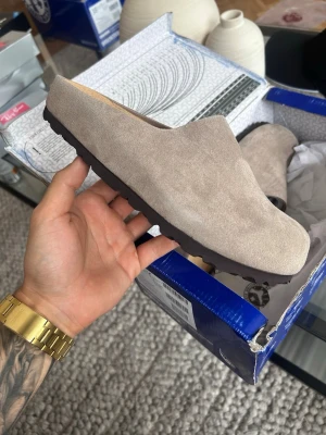 Beige mockatofflor från Birkenstock - Snygga beige tofflor från Birkenstock i mjuk mocka med klassisk slip-in design och mörk sula. Perfekta för dig som vill ha både stil och komfort hemma. Tidlös look som passar till det mesta.
