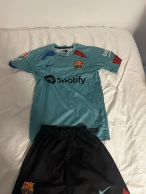Barcelona bortaställ Gavi #6 Nike - Säljer ett Barcelona bortaställ med Gavi #6. Tröjan är ljusblå med klubbmärke, Nike-logga och Spotify som sponsor. Färgdetaljer i rött och blått på ärmarna. Tillhörande svarta shorts med klubbmärke och Nike-logga. Perfekt för fotbollsträning eller match.