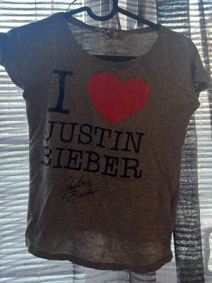Grå I Love Justin Bieber t-shirt - Säljer en grå t-shirt med trycket 'I ♥ Justin Bieber' i svart och rött framtill. T-shirten har rund halsringning och korta ärmar. Perfekt för dig som är ett fan av Justin Bieber och vill visa det med stil.