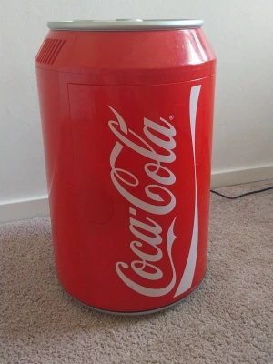 Coca-Cola minikyl i burkdesign - Säljer en minikyl som ser ut som en stor Coca-Cola burk. Kylen är röd med klassisk Coca-Cola logga och har ett hyllplan inuti. Perfekt för att förvara drycker eller snacks på rummet eller kontoret.