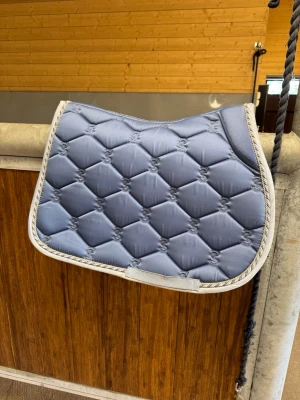 Blått schabrak från PS of Sweden - Säljer ett snyggt blått schabrak från PS of Sweden med quiltat mönster och broderad text längs kanten. Schabraket har ljus kant och är perfekt till ridning. Materialet är glansigt och ger en exklusiv känsla. Storlek ponny och bara testat. Annnars helt nytt 