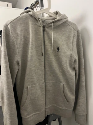 Ljusgrå hoodie från Polo Ralph Lauren - Snygg ljusgrå hoodie med dragkedja från Polo Ralph Lauren. Klassisk design med huva och vita dragsnören, två fickor framtill och den ikoniska loggan broderad på bröstet. Tillverkad i mjukt material som känns skönt mot huden. Storlek Xs Passae S också 
