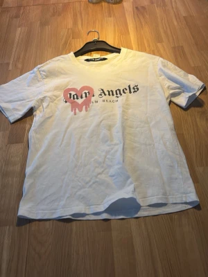 Vit t-shirt med tryck Palm Angels - Vit t-shirt med svart och rosa tryck på bröstet där det står 'Palm Angels Palm Beach'. Klassisk passform med korta ärmar och rund halsringning. Perfekt för en avslappnad och trendig stil.