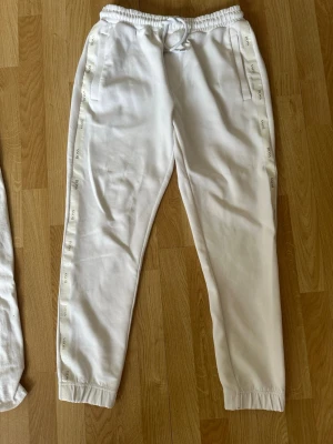 Vita Hugo BOSS mjukisbyxor - Säljer ett par vita mjukisbyxor från Hugo Boss med resår i midjan och benslut. Byxorna har sidofickor och ett dekorativt band med logga längs båda benen. Sköna och avslappnade, perfekta för chill dagar. Materialet känns mjukt och stretchigt.