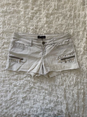 Coola vita lågmidjade jeansshorts  - Snygga vita denimshorts från Melrose i storlek 38 med unika detaljer! Midjemåttet är 39cm. Modellen har en rå, fransig kant nertill och är dekorerad med dekorativa dragkedjor samt glittriga nitar och strassdetaljer både fram och på ena bakfickan. Perfekta till sommarens alla outfits och festivaler. I fint skick – hör av dig vid frågor! ✨