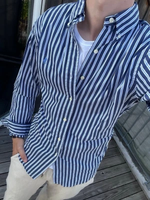Blåvit randig skjorta Polo Ralph Lauren - Snygg blå skjorta med vita ränder från Polo Ralph Lauren. Skjortan har klassisk krage, knappar framtill och lång ärm. Custom fit-modell som sitter riktigt bra och passar till många olika stilar.