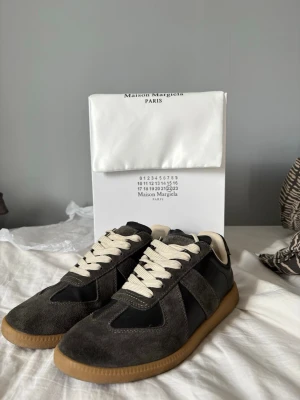 Maison Margiela svarta sneakers - Maison Margiela sneakers i svart mocka och läder med beigea skosnören och klassisk gummisula. Snygg retrostil med låg profil och diskreta detaljer. Perfekta för dig som gillar minimalistisk och stilren design.