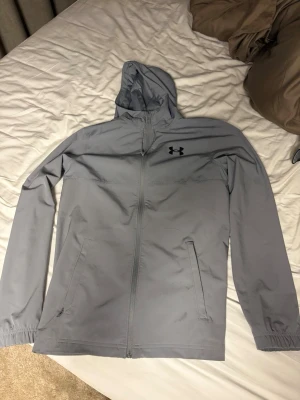 Grå vindjacka från Under Armour - Snygg grå vindjacka från Under Armour med huva och hel dragkedja framtill. Jackan har två sidofickor med dragkedja och elastiska muddar vid ärmsluten. Under Armour-logga på bröstet och stor text på ryggen. Perfekt för blåsiga dagar.