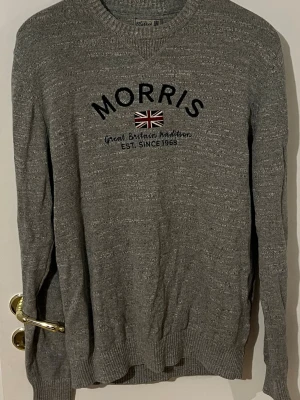Grå stickad Morris tröja - Snygg gråmelerad stickad tröja från Morris med broderad text och brittisk flagga på bröstet. Rund halsringning och ribbade muddar vid ärmslut och nederkant. Perfekt för dig som gillar klassisk och stilren design. Pris kan diskuteras vid snabb affär