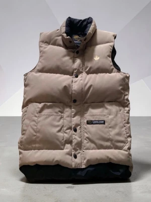 Beige dunväst från Canada Goose - Säljer en beige dunväst från Canada Goose med svart innerfoder och tryckknappar framtill. Västen har två stora framfickor och en broderad logga på bröstet. Perfekt för lager-på-lager och riktigt snygg till höst- och vinterlooken.