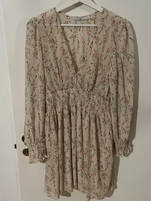 Blommig kortklänning med v-ringning - Söt kortklänning i beige med blommigt mönster i rosa och grönt. Klänningen har v-ringning, långa ärmar med smock vid ärmslut och markerad midja med smock. Perfekt för sommarens alla tillfällen! köpt för 699:- Andvänd 1 gång men säljes för den ej andvänds. Pris kan diskuteras vid snabb affär.