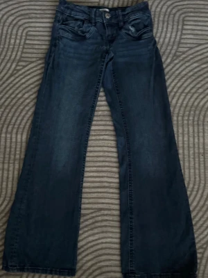 Mörkblå bootcut jeans - Priset går att diskuteras. Snygga mörkblå jeans med bootcut passform och klassiska fem fickor. Jeansen har en lätt tvättad look och raka ben som blir vidare nedtill. Perfekta för dig som gillar en avslappnad men trendig stil.