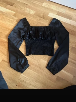 Svart croppad topp från FB Sister - Säljer en svart croppad topp från FB Sister med långa puffärmar och smockad midja. Toppen har en snygg volangdetalj upptill och är tillverkad i ett glansigt material som ger en lyxig känsla. Perfekt för dig som vill sticka ut lite extra.