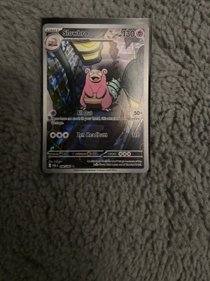 Pokémonkort Slowbro holo - Säljer ett Pokémonkort med motivet Slowbro. Kortet har en holografisk yta och visar Slowbro i rosa färg med en stadsmiljö i bakgrunden. Text och effekter syns tydligt på framsidan. Perfekt för samlare eller Pokémonfans.