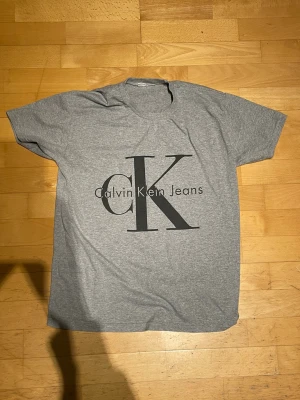Grå Calvin Klein Jeans t-shirt - Snygg grå t-shirt från Calvin Klein Jeans med stort CK-tryck på bröstet. Klassisk passform och rund halsringning. Perfekt till jeans eller shorts för en clean och enkel look.