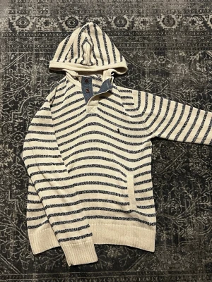 ralph lauren hoodie  - Säljer en beige och mörkblå randig hoodie från Ralph lauren i stickad bomull. Hoodien har huva med dragsko och snygga träknappar vid halsen. Ribbstickade muddar vid ärmslut och nederkant. Perfekt för dig som gillar en avslappnad och stilren look. Använd en gång, den är helt ny 