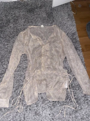 Beige blommig blus med knytband - Säljer en tunn och luftig beige blus med diskret blommönster och knytband i midjan. Blusen har krage, knäppning framtill och långa ärmar. Perfekt för en trendig och avslappnad look.